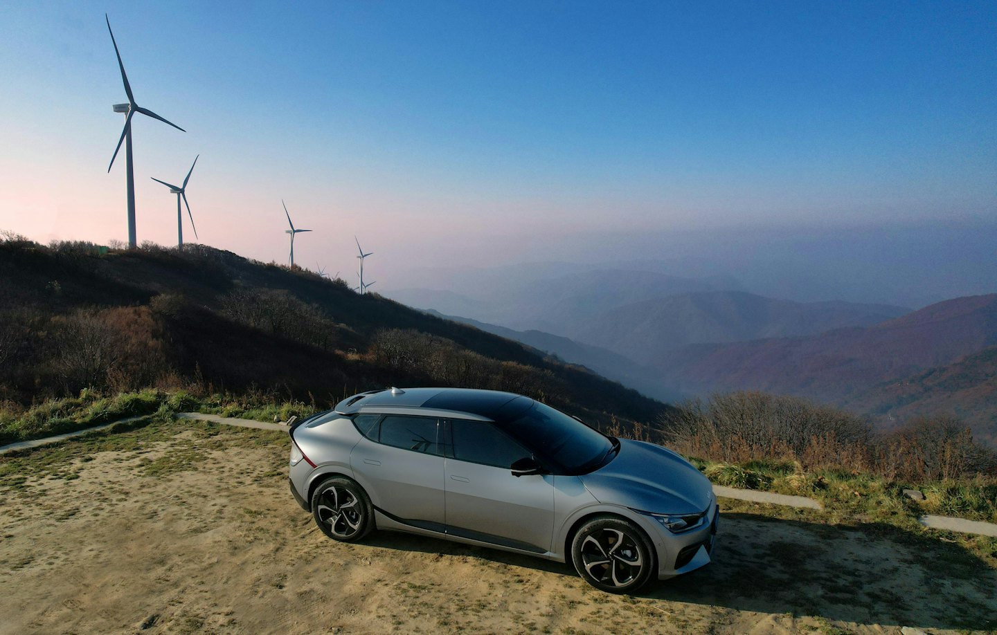 Silver electric SUV parked on dirt path with wind turbines.